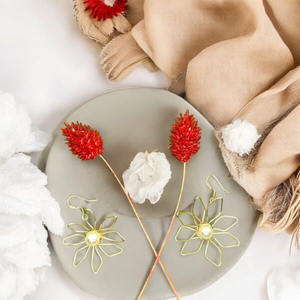 Pendientes Zoe con diseño floral de 8 pétalos en alambre dorado y perla central, sobre un plato decorativo con flores rojas y beige.