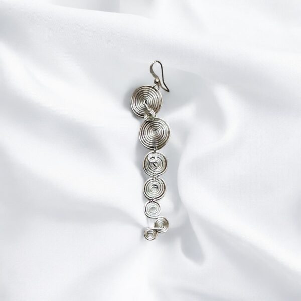 Pendientes Largos Espiral You formado por distintos tamaños de círculos plateados sobre fondo de tela blanca.